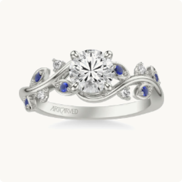 Frida. ArtCarved Sapphire & Diamond Petal Accent Setting in 14k White Gold: Round-cut diamond ring surrounded by a vine-like white gold band with sapphire and diamond leaf accents, engraved with the ArtCarved name.