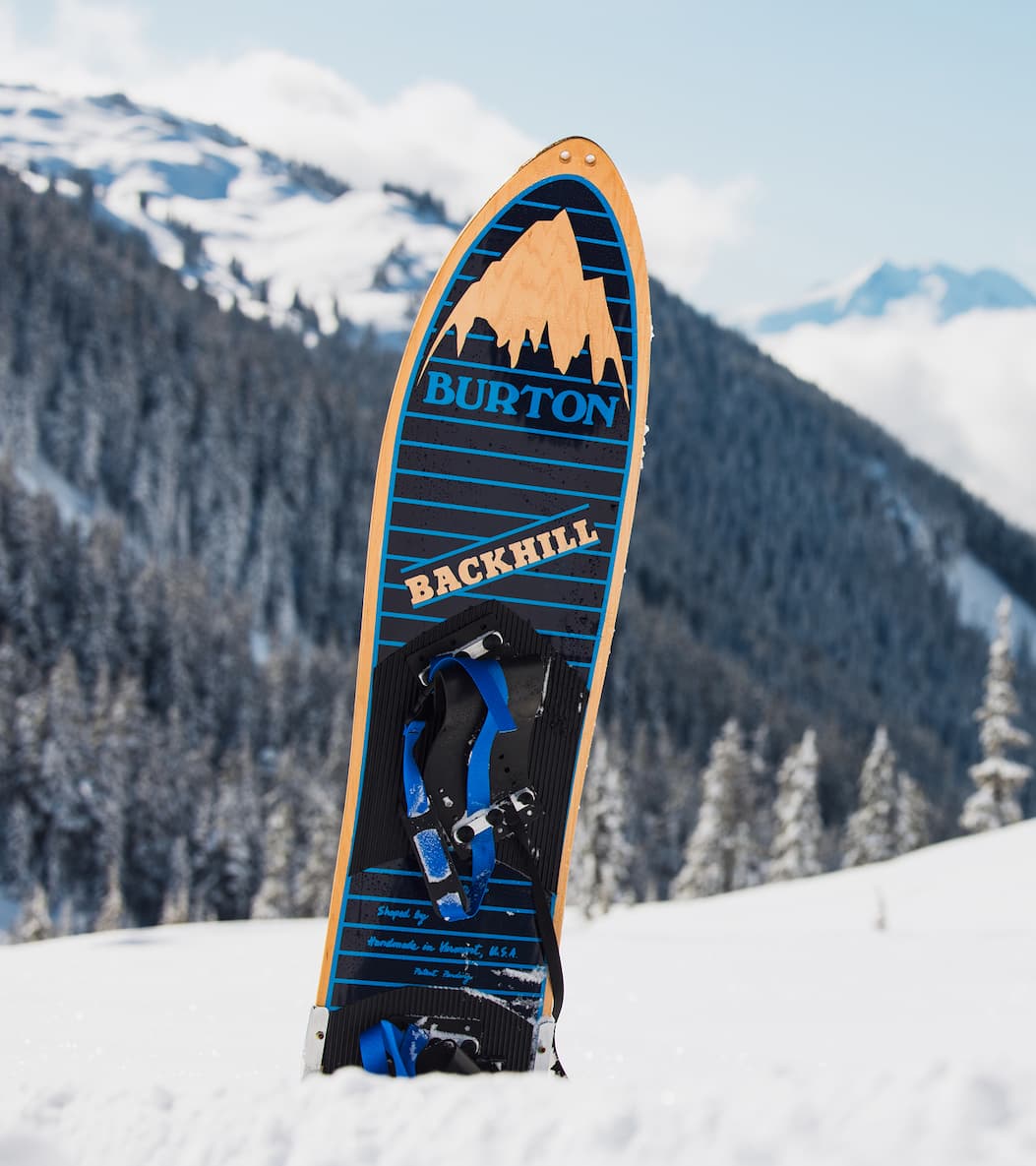 1985 Burton Backhill snowboard with mountain graphic, standing upright in snow with forested mountains in the background.