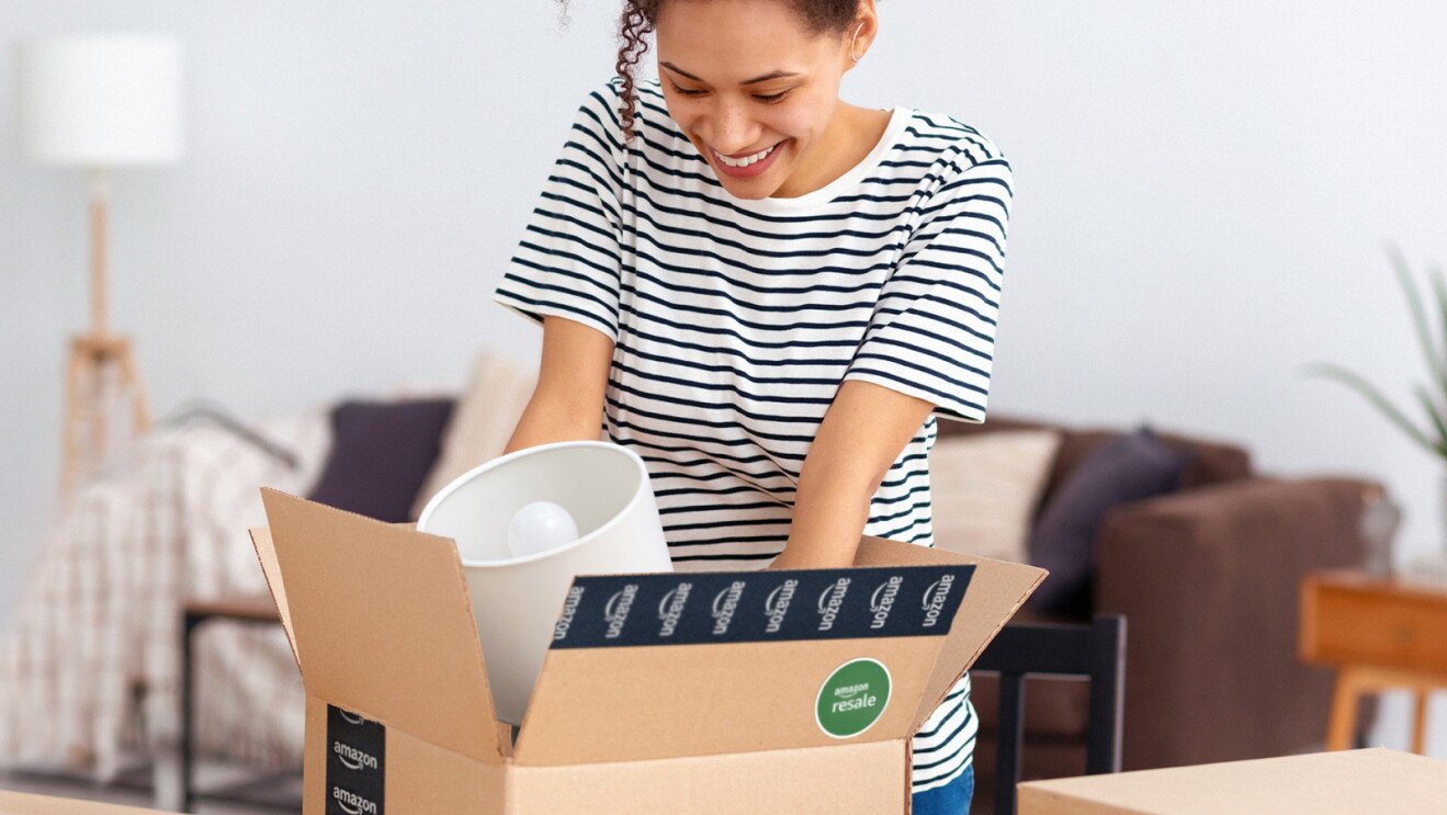 A woman in a striped shirt opens an Amazon Resale box and lifts out a lamp