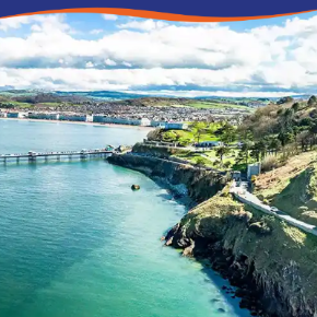 Aerial view of a coastal landscape featuring cliffs, a pier, and lush greenery under a blue sky.