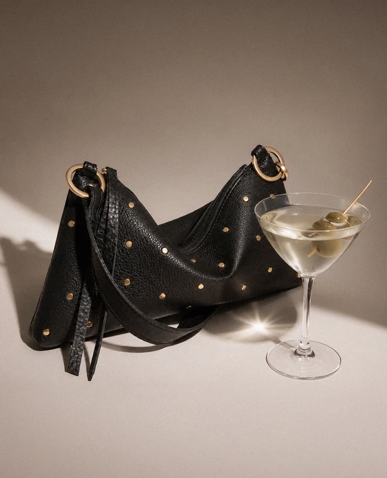 Kyle Sml in Starlight Black leather shoulder bag with mini brushed gold rivets styled beside a martini glass with olives.