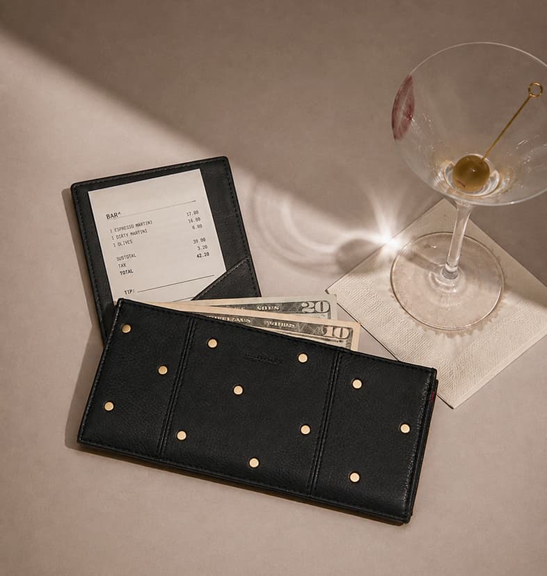110 Noth Starlight Black leather wallet with mini brushed gold rivets, styled with cash, a restaurant check, and a martini glass.