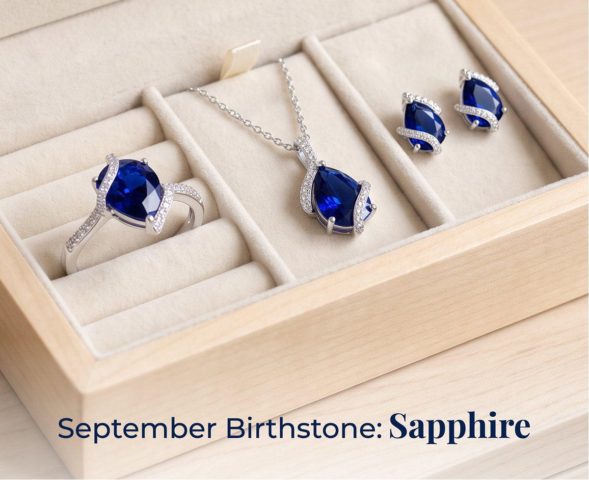 Sapphire and diamond jewelry set — a ring, pendant necklace, and stud earrings, each featuring a blue sapphire wrapped in a diamond swirl accent — displayed in a light wood jewelry box, with text reading 'September Birthstone: Sapphire.'