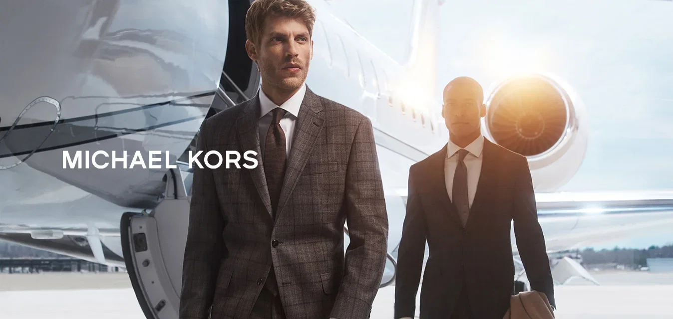 Twee mannen in op maat gemaakte pakken lopen langs een privéjet. De eerste man draagt een bruine leren tas, terwijl de tweede een beige overjas vasthoudt.