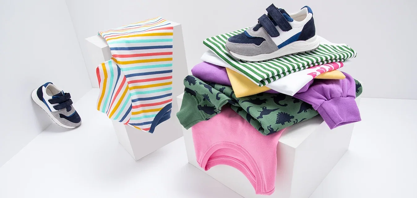 Kindersneakers in marineblauw, grijs en wit, naast opgevouwen kleding: gestreepte, groene shirts met dinosaurusprint, paarse, roze, gele en witte shirts.