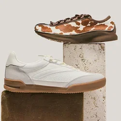 Twee casual sneakers getoond op gestapelde rechthoekige blokken; één wit met een beige zool, de andere bruin met een wit koeienprintpatroon.