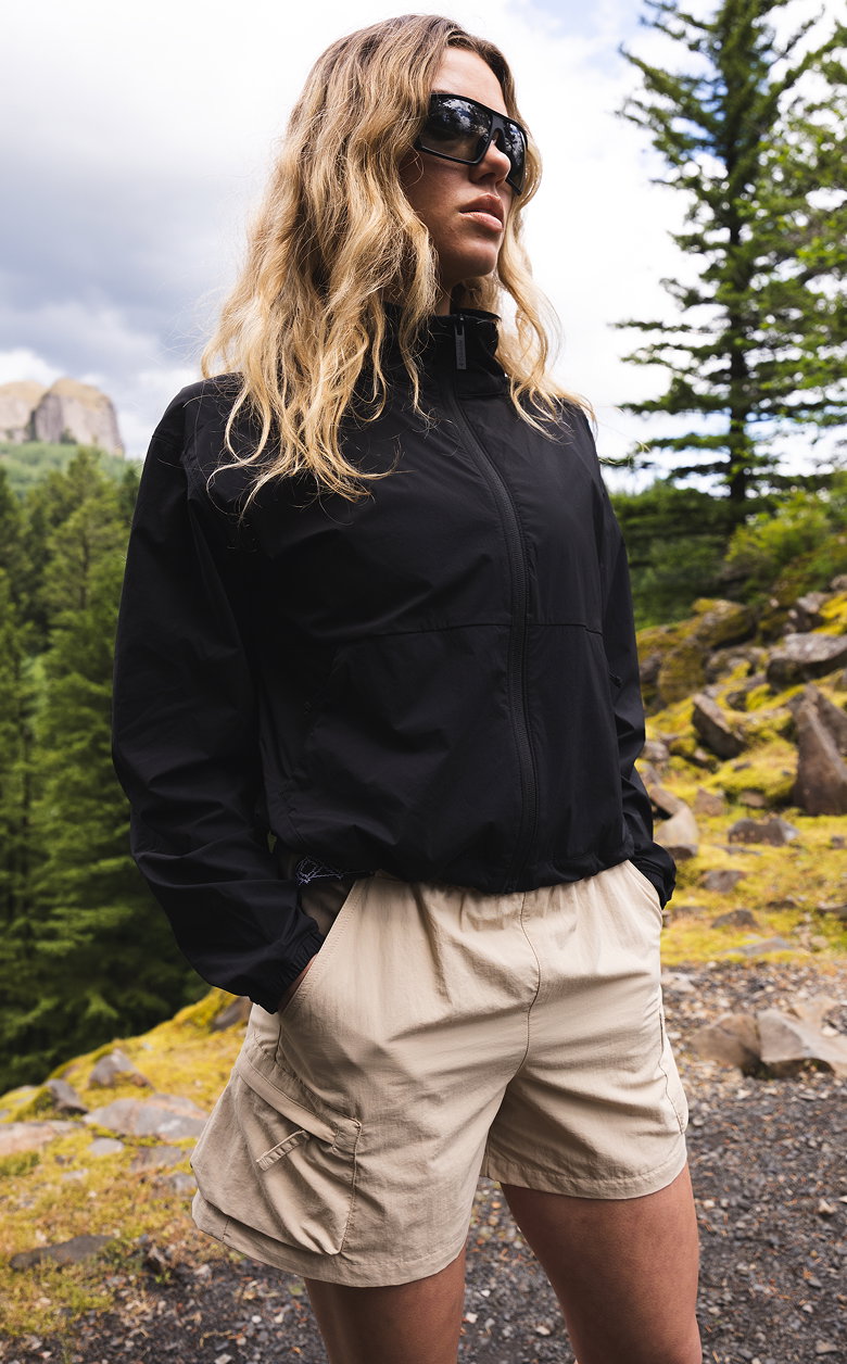 A woman standing on a hilltop wearing a black jacket and beige shorts.