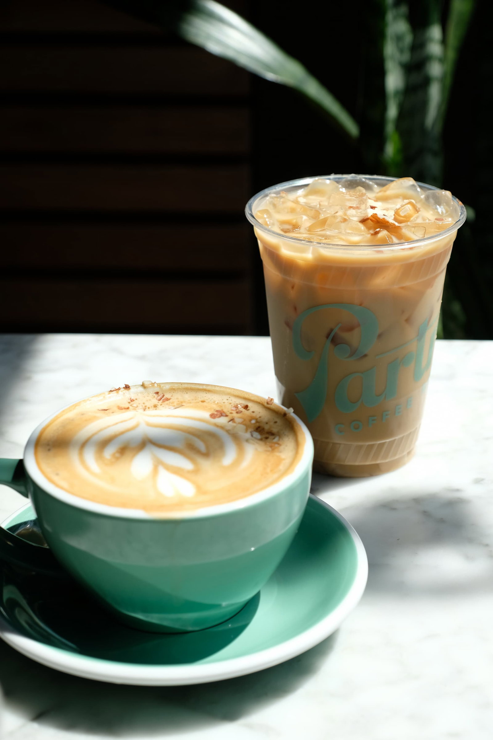 A hot Almond Maple Latte in a mug next to an iced Almond Maple Latte in a Partners Coffee cup