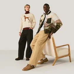 Twee modellen poseren in een neutrale ruimte; de een staat in een hondentrui en zwarte broek, de ander leunt op een houten fauteuil, gekleed in een patroonjas en beige broek.