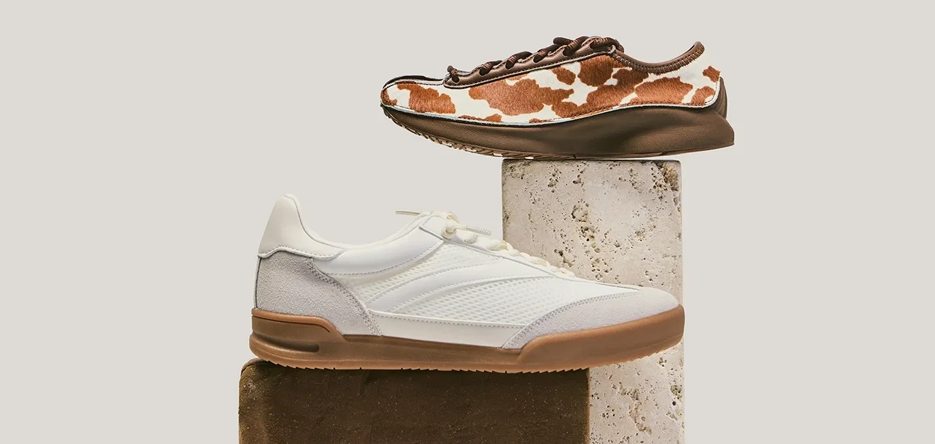 Twee casual sneakers getoond op gestapelde rechthoekige blokken; één wit met een beige zool, de andere bruin met een wit koeienprintpatroon.