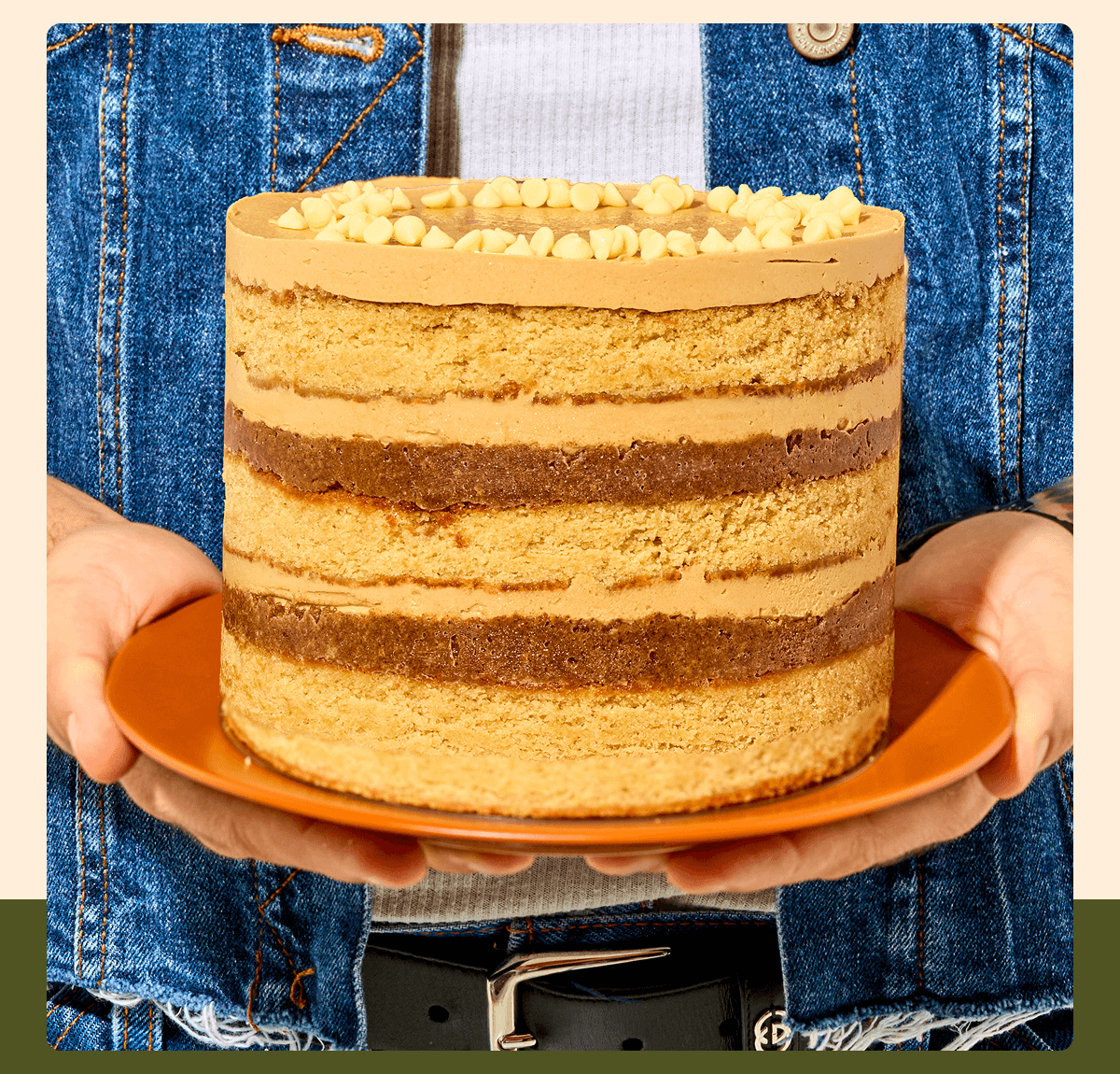 Person in a denim vest holds an orange plate with a 6-inch Maple Cookie Butter Cake, featuring layers of cake, cookie butter frosting, crunchy filling, and white chocolate chips on top.