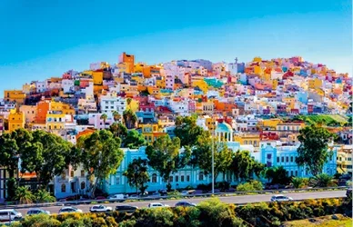 Colourful hillside cityscape with vibrant buildings, trees and coastal road under a bright blue sky.