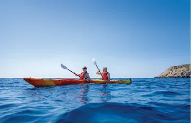 Two people kayaking across clear blue sea near a rocky coastline.