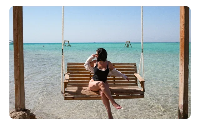 A woman in a black bikini on a wooden swing over shallow turquoise water, weathered brown posts framing scene, natural rope, clear aqua sea, sandy bottom visible, distant figures, pale blue sky and tropical beach setting.