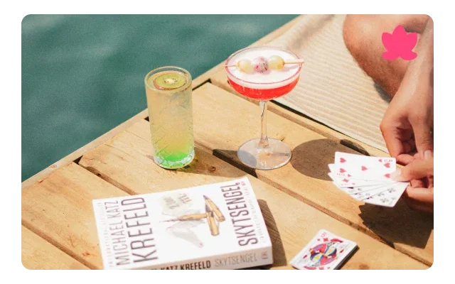 Overhead view, turquoise pool water, light wood deck, two cocktails in tall glass and coupe glass with red rim, book titled 'Reckless' by Miranda Krefeld, hand holding playing cards with hearts, pink flower accent and bright sunlight.
