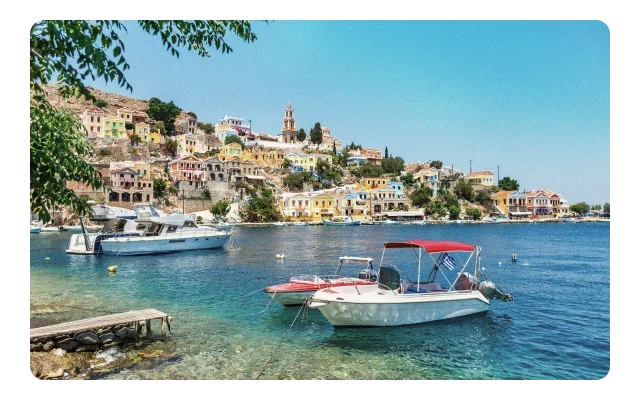 Mediterranean coastal village, pastel buildings on hillside, church bell tower with dome, turquoise water, white motorboat with blue canopy, wooden dock, white yacht, green cypress trees, clear blue sky and Greek island architecture.