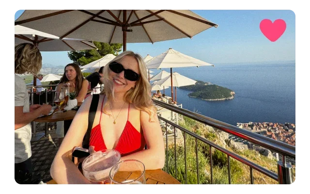 Blonde woman in red bikini top, black sunglasses, gold necklace, holding clear cup, smiling, terrace with beige umbrellas, black railing, Adriatic Sea, forested island, terracotta roofs, blue sky and a pink heart icon overlay.