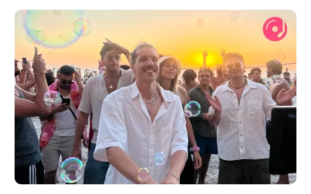 Person in white shirt, silver necklace, smiling, golden hour sunset, peachy-orange sky, crowd with raised phones, white clothing, iridescent soap bubble overlays, pink, turquoise, white bubbles, rainbow reflections, backlit silhouettes and festive outdoor gathering.