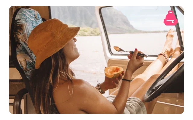 Woman in tan bucket hat eating grapefruit with spoon inside vehicle, brown hair, cream top, patterned seat, black steering wheel, lake view through windows, misty mountains, overcast sky, pink van icon overlay and warm interior tones.