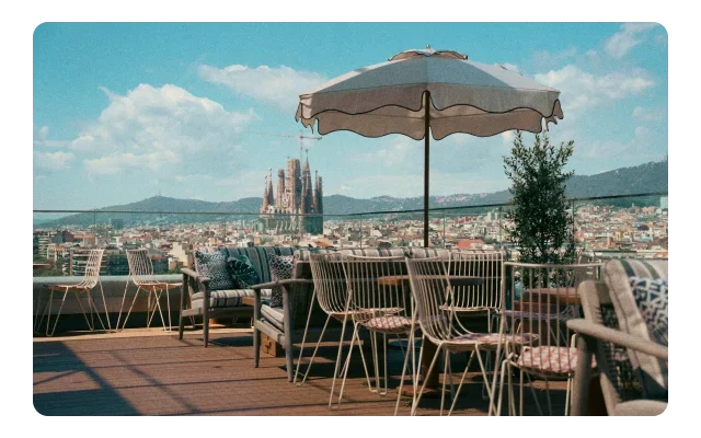 Rooftop terrace with wooden deck, round table, rose gold wire chairs, beige fringed umbrella, teal sofa, glass railing, Barcelona cityscape, Sagrada Familia basilica, beige buildings, blue-grey mountains, turquoise sky and white clouds.