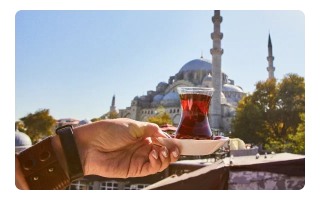 Hand holding traditional Turkish tulip-shaped tea glass with amber-red drink in a cream saucer, dark railing, green foliage, Ottoman mosque with blue-grey domes, two minarets, pale blue sky and Istanbul cityscape.