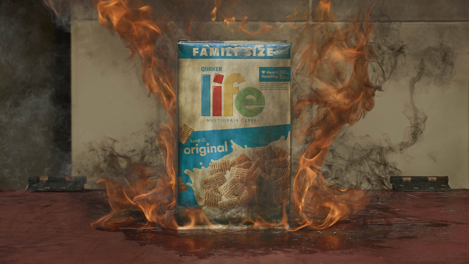 A family-size box of Life cereal stands upright on a flat surface, engulfed in orange flames and dark smoke.