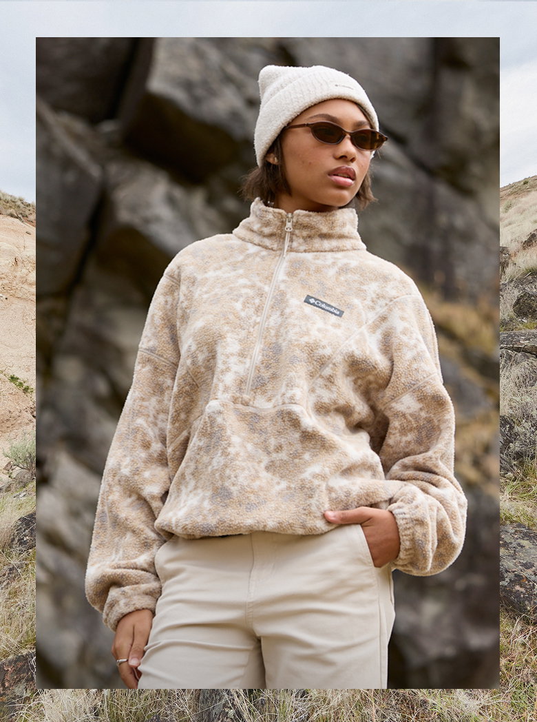 A woman in a tan fleece jacket and white beanie standing in a rocky landscape.