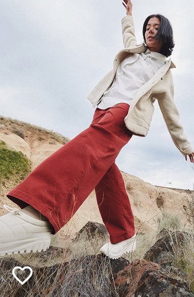 A woman wearing a white jacket and red pants as she walks outside over rocks.
