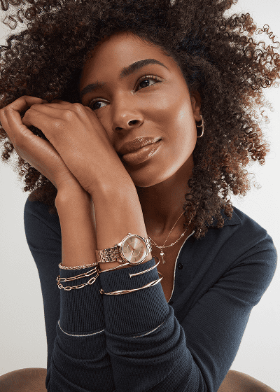 Woman wearing layered yellow gold jewelry, including bracelets, a watch, necklaces and hoop earrings.