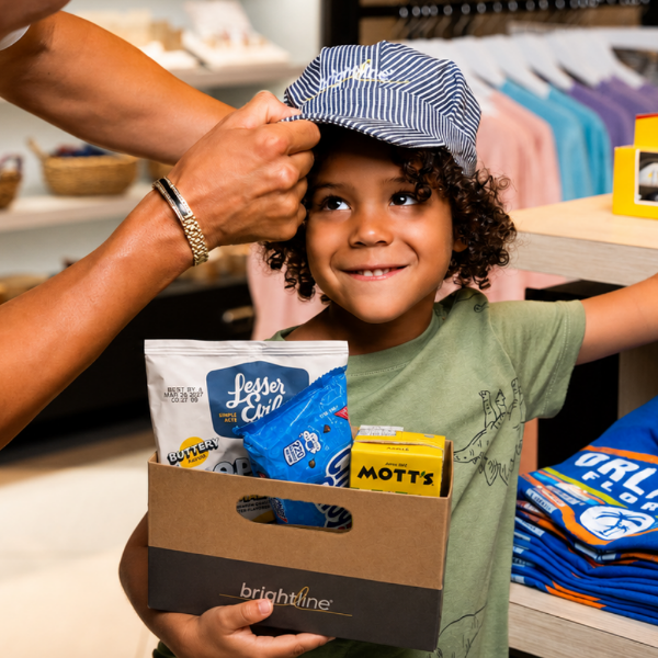 A young child smiles while an adult adjusts a striped cap on their head. The child holds a cardboard snack box containing items including a LesserEvil snack bag and a Mott's product. They stand inside a brightly lit retail environment with folded clothing and other merchandise visible in the background. The word "brightline" appears printed on the front of the box.