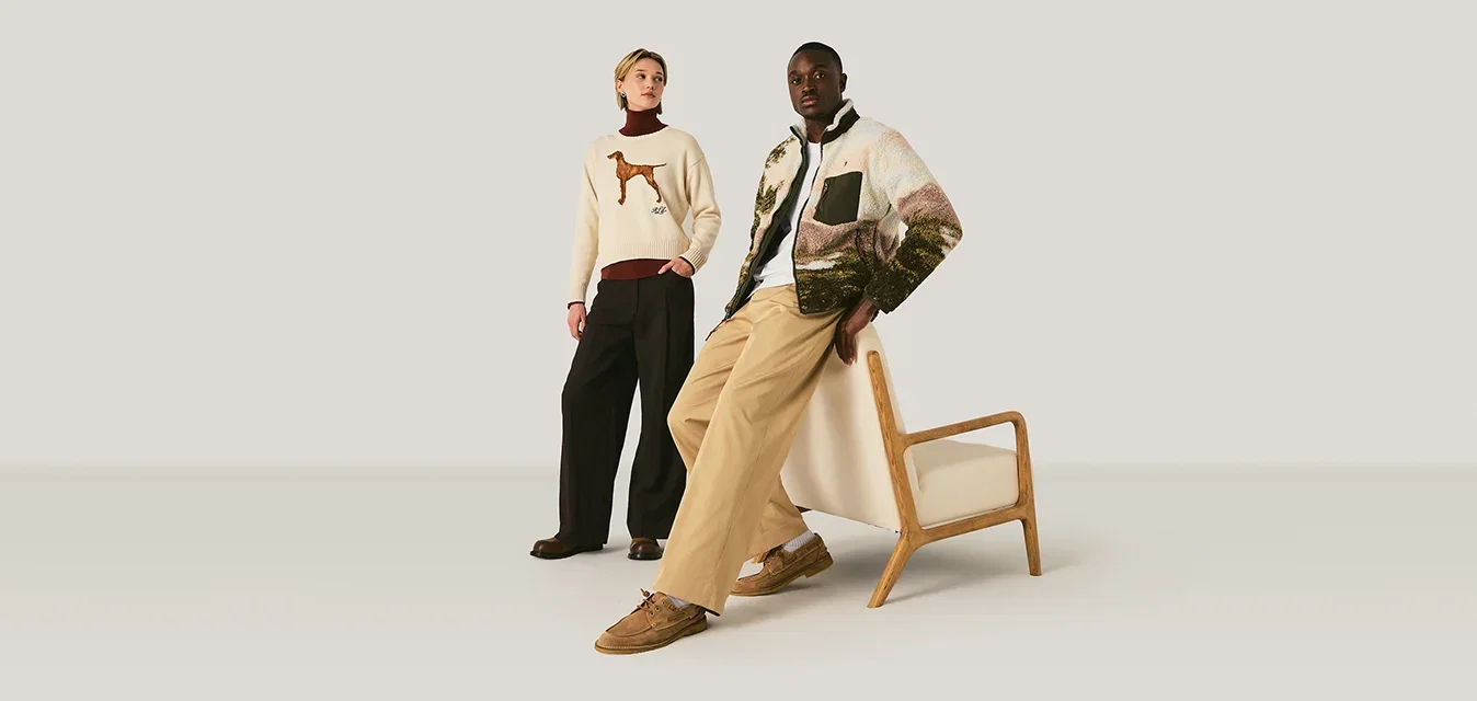 Twee modellen poseren in een neutrale ruimte; de een staat in een hondentrui en zwarte broek, de ander leunt op een houten fauteuil, gekleed in een patroonjas en beige broek.