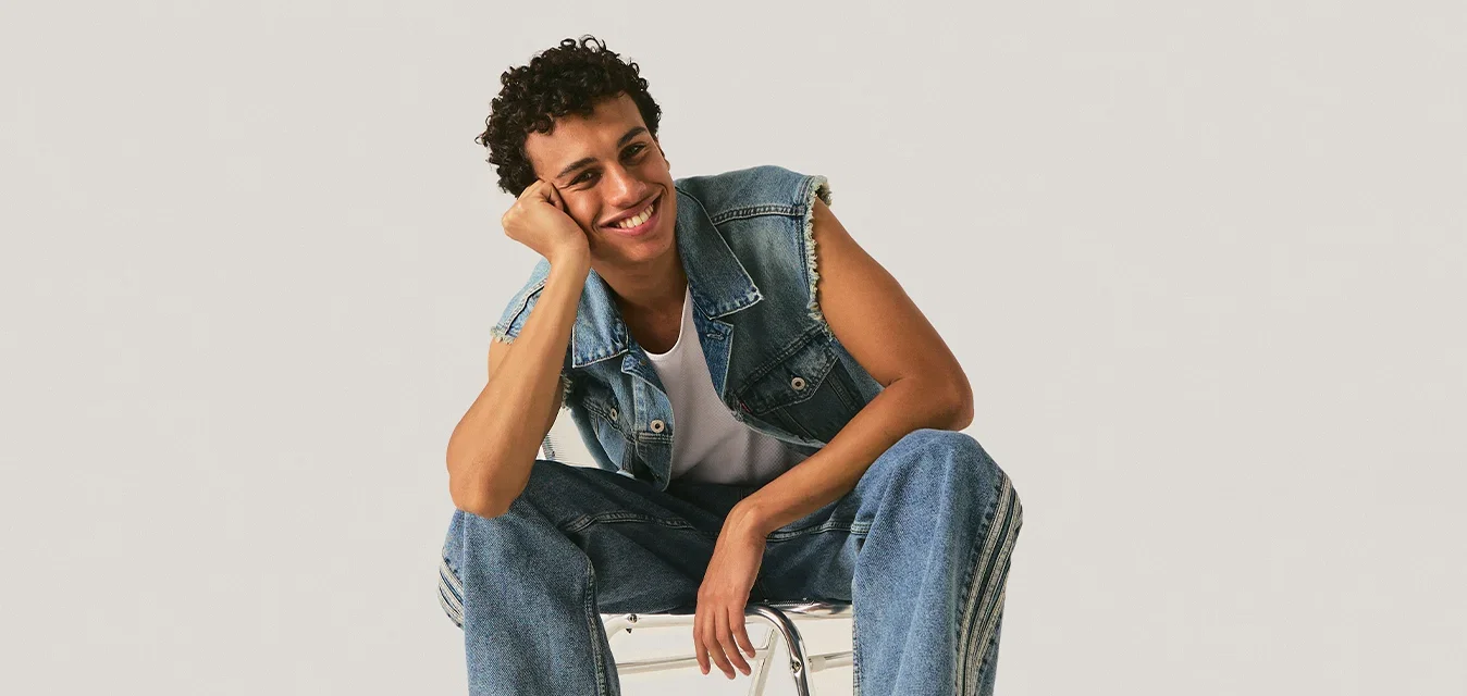 Jong persoon zittend op een minimalistische stoel, draagt een denim vest en jeans, zwarte laarzen, glimlachend, met een eenvoudige lichte achtergrond.