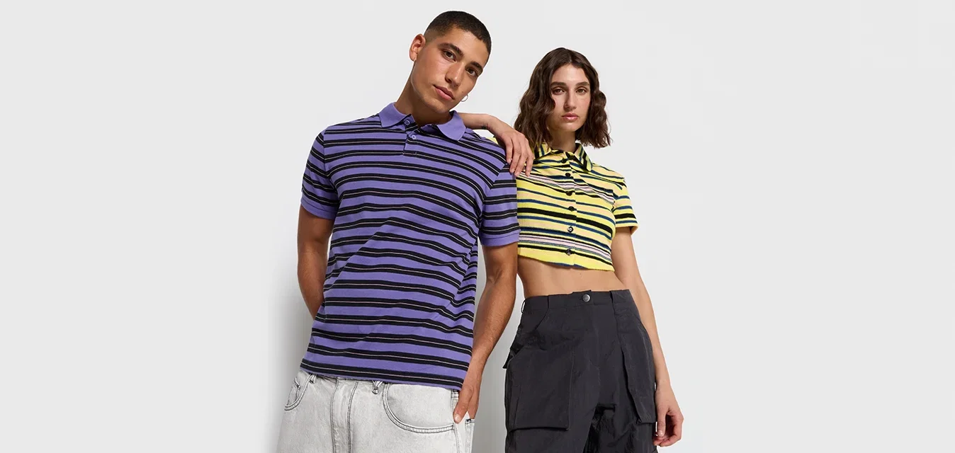 Twee personen dragen gestreepte polo's; de man in paars en zwart, en de vrouw in geel en zwart. Beide outfits hebben korte mouwen en een casual pasvorm.