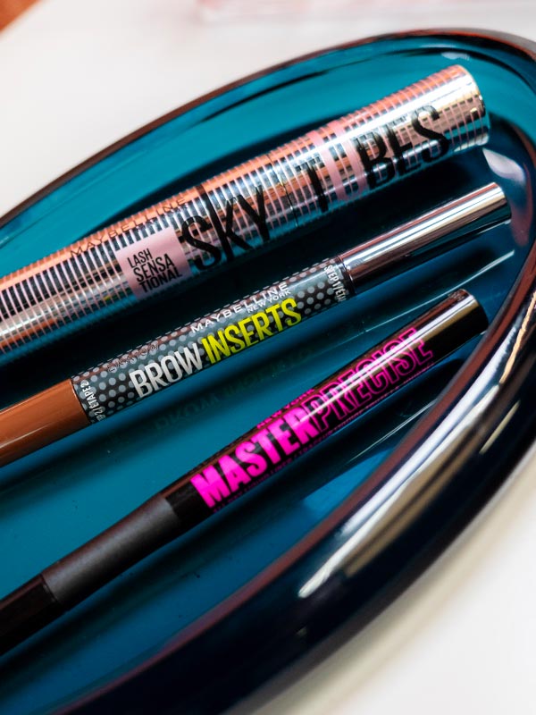 A lifestyle image of Maybelline eye makeup trio for The Makeup Relationship Test Challenge #2: Eyes, featuring Sky Tubes Tubing Mascara, Brow Inserts Multiplying Tool, and Master Precise Liquid Eyeliner arranged on a glossy teal tray.
