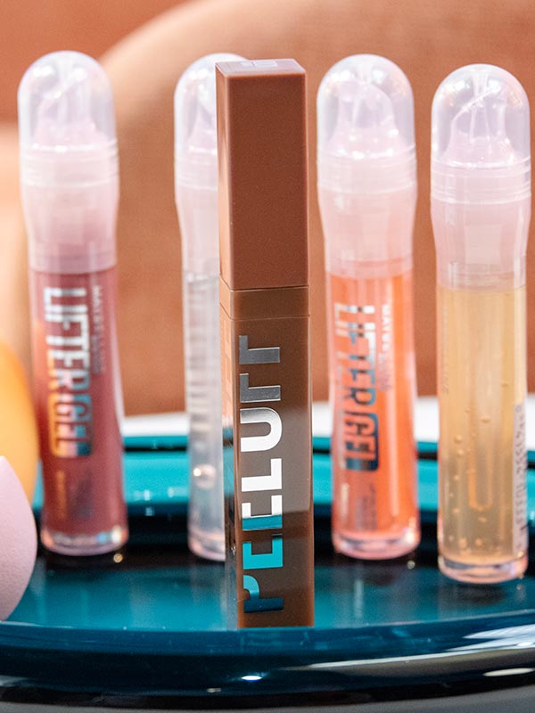 A lifestyle image of Maybelline lip lineup for The Makeup Relationship Test Challenge #3: Lips, featuring a brown Super Stay Peel-Off Lip Liner with several different shades of Lifter Gel Lip Gloss behind it arranged on a glossy teal tray.
