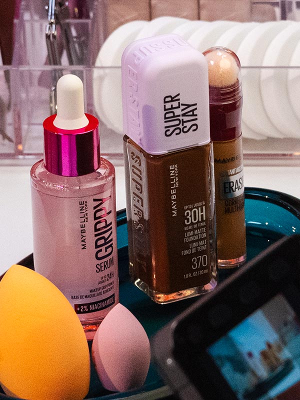 A lifestyle image of a Maybelline face makeup lineup for The Makeup Relationship Test Challenge #1: Face, featuring Grippy Serum Grip Primer, Super Stay Lumi-Matte Foundation, Instant Eraser Concealer, and two blending sponges arranged on a glossy teal tray.