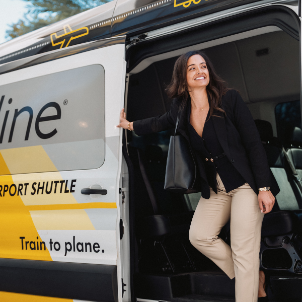  A smiling woman steps out of a modern airport shuttle van. She is dressed in business attire with a blazer, trousers, and a shoulder bag, suggesting a professional trip. The shuttle features a bright yellow and white design with text indicating it connects train to plane. The interior of the van is visible behind her, emphasizing comfortable group transportation.