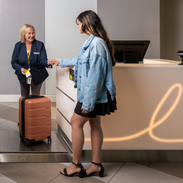 A young woman hands an item to a smiling receptionist at a modern front desk. The traveler stands beside a brown rolling suitcase placed on a platform near the counter. The receptionist wears a navy blazer and name badge, suggesting a professional hospitality setting. Soft lighting and neutral tones create a welcoming, contemporary atmosphere.