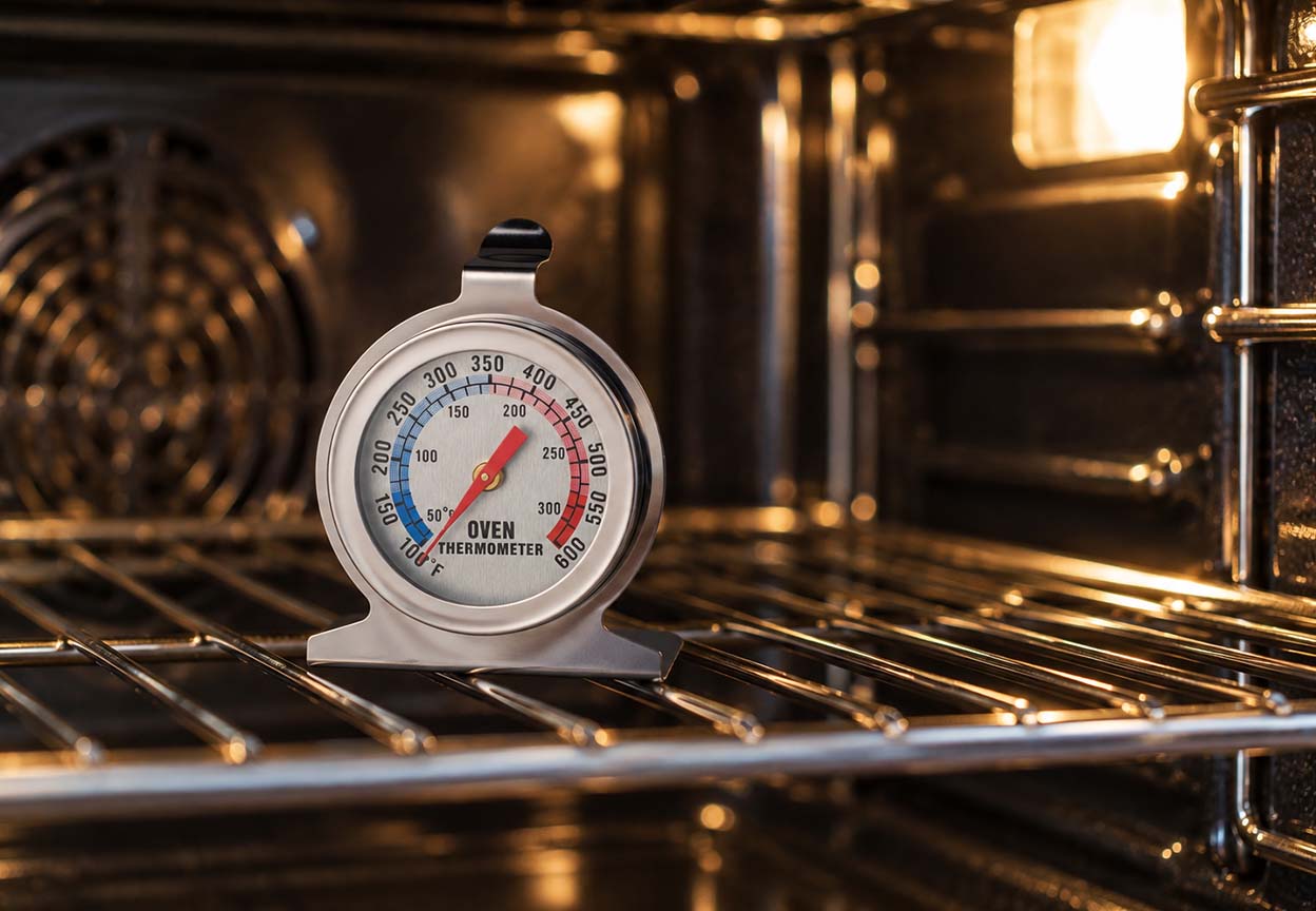 Analog oven thermometer rests on a rack inside a warmly lit oven