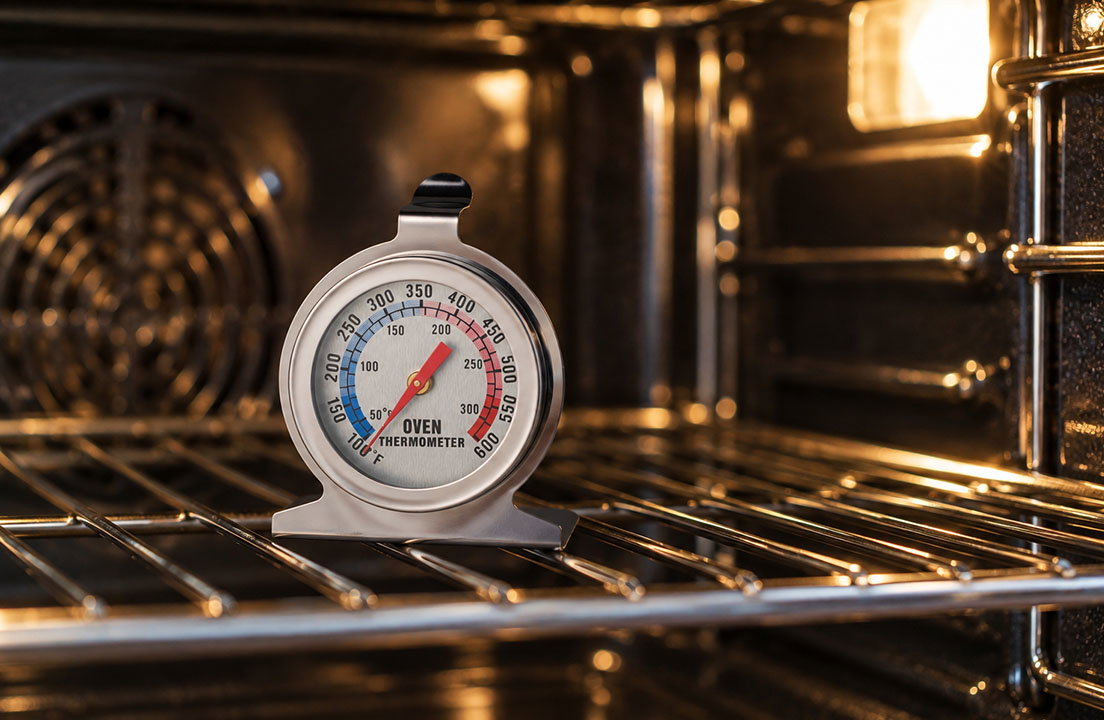 Analog oven thermometer rests on a rack inside a warmly lit oven