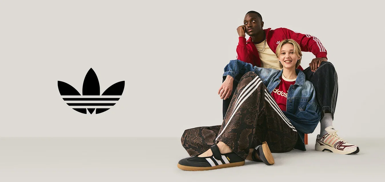 Een man in een rode jas en een vrouw in een spijkerjasje en Adidas-broek zitten op de vloer tegen een effen lichte achtergrond.