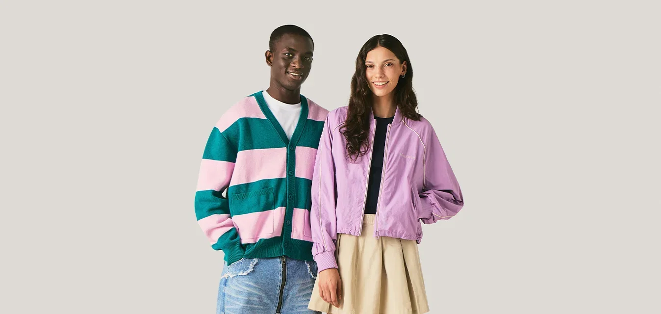 Lachende jonge man in groen-roze gestreepte trui staat naast lachende jonge vrouw in paarse jas en beige rok tegen een effen achtergrond.