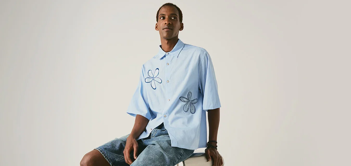 Jonge man zit op een kruk, draagt een lichtblauw shirt met bloemenprint en spijkershorts, tegen een effen lichte achtergrond.
