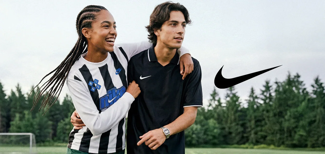 Twee jonge mensen lopen op een voetbalveld, de een met zijn arm om de ander, beiden dragen sportkleding en lachen.