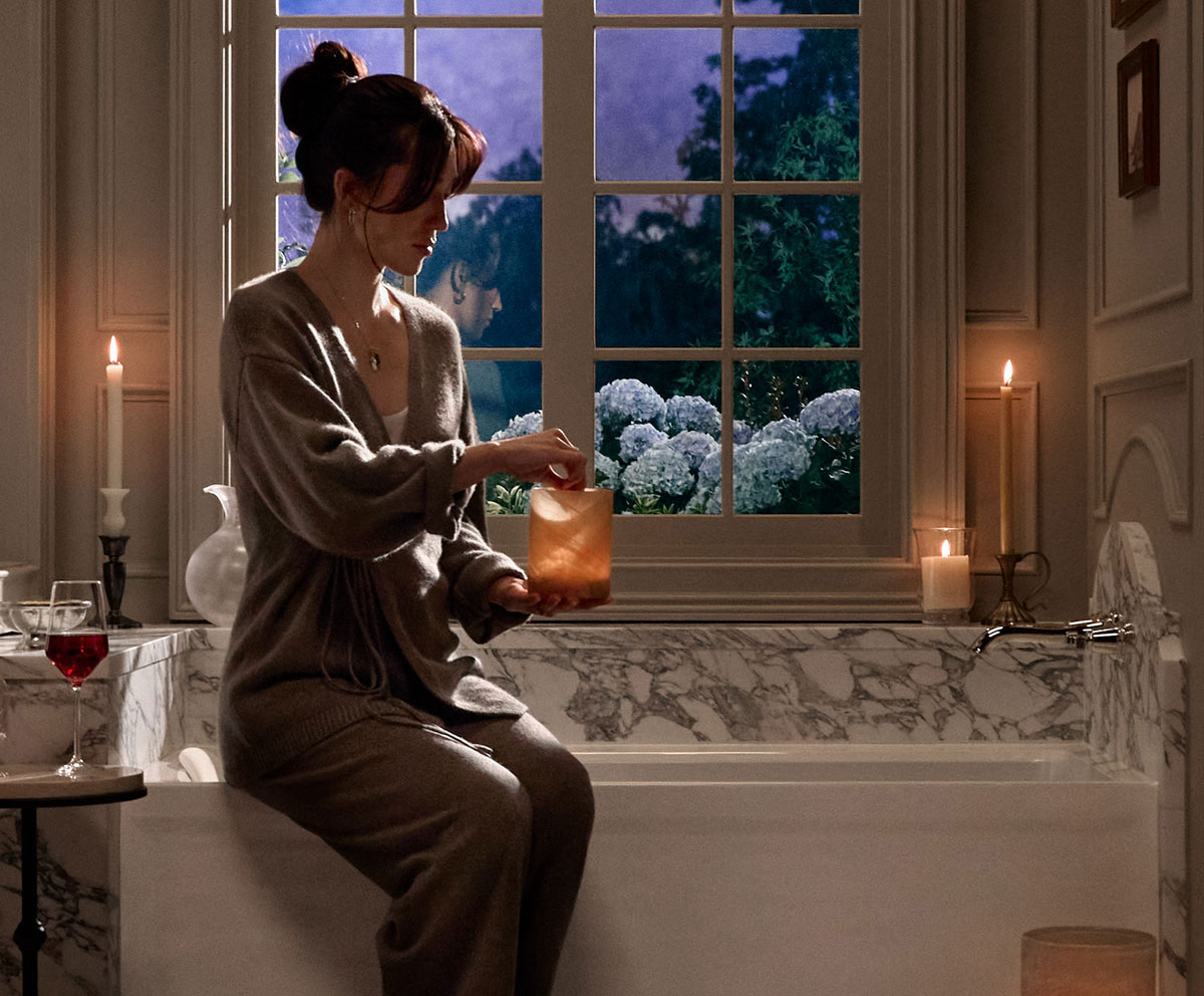 Person seated on the edge of an alcove bath  in a softly lit bathroom, lighting a candle across from a   large window.