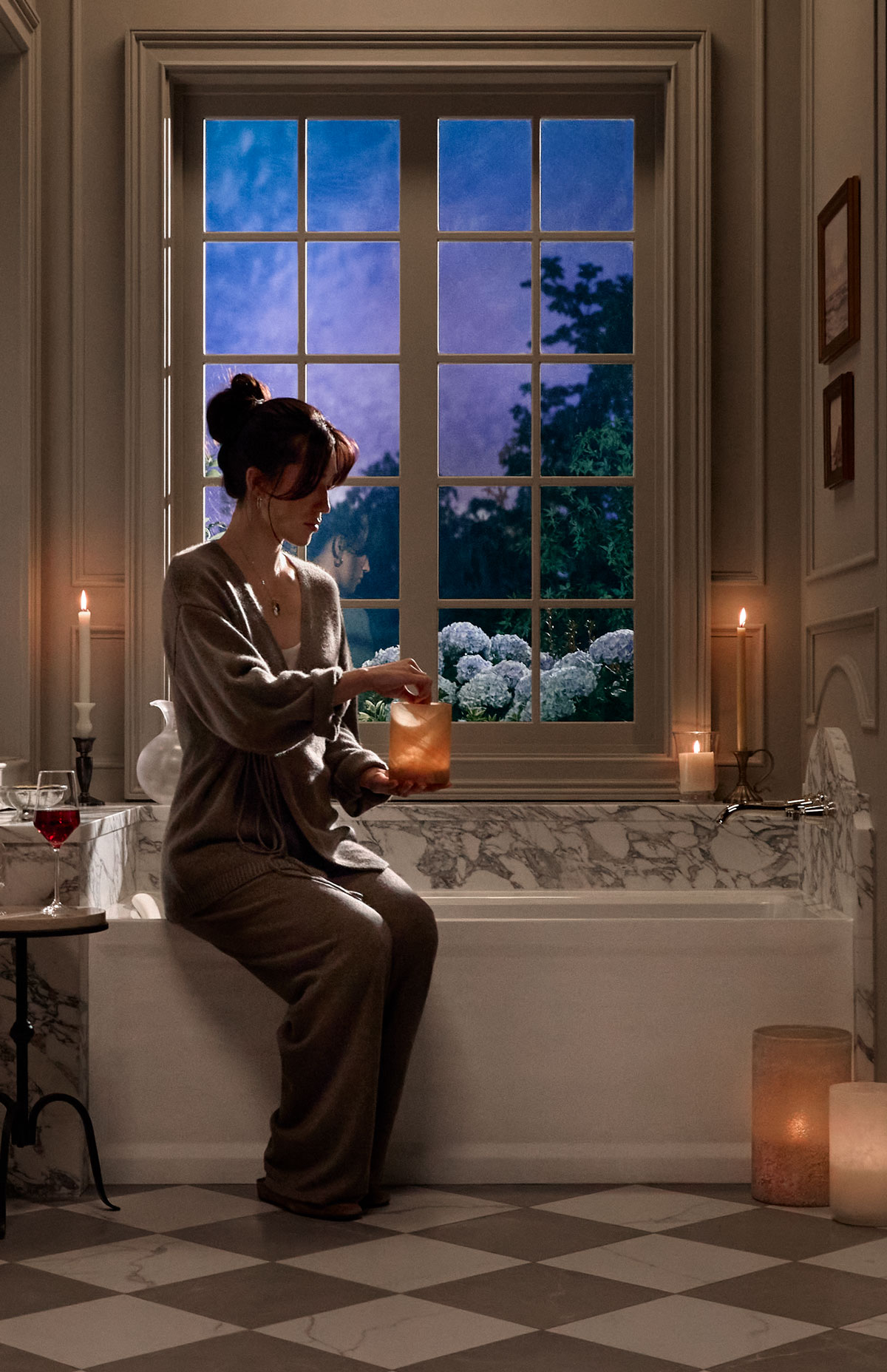 Person seated on the edge of an alcove bath  in a softly lit bathroom, lighting a candle across from a   large window.
