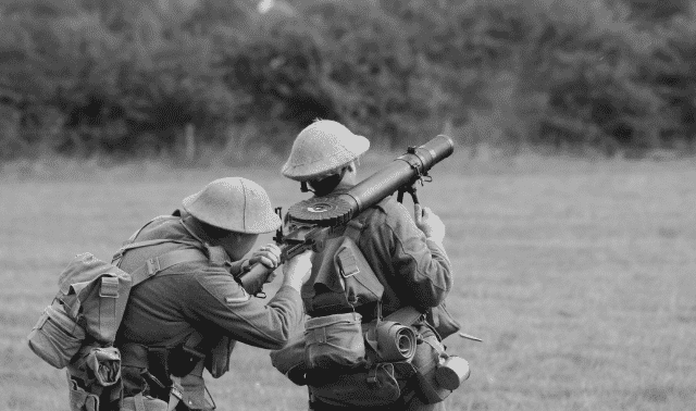 Two soldiers in vintage military uniforms and helmets operate an old-fashioned, mounted machine gun in a grassy field with trees in the background.