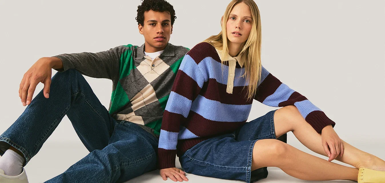 Twee jonge volwassenen zitten op de vloer tegen een effen achtergrond, gekleed in casual sweaters, denim broeken en neutraal schoeisel.