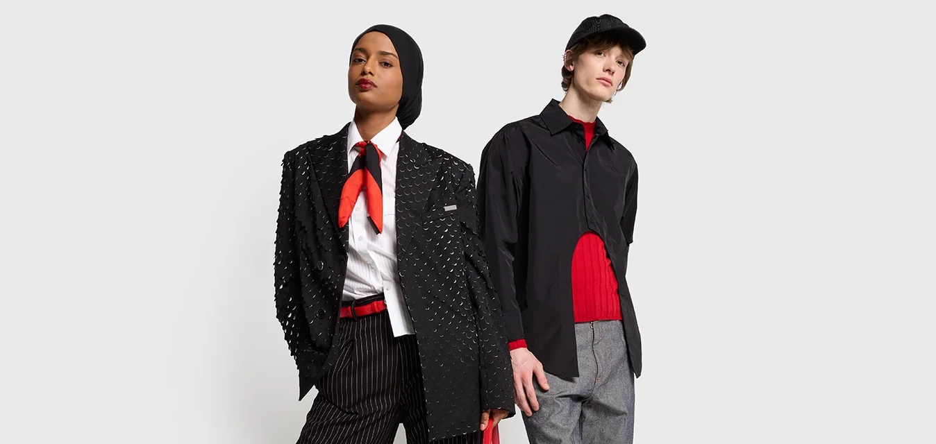 Twee modellen staan zij aan zij. Het vrouwelijke model draagt een gestructureerde zwarte blazer, gestreepte broek en rode accenten. Het mannelijke model draagt een zwart shirt, een rode trui en een grijze broek.