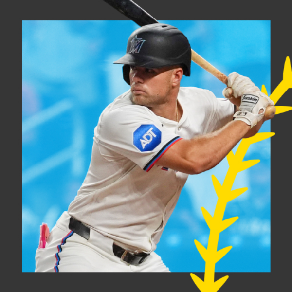 A baseball player in a white uniform prepares to swing the bat during a game. He wears a black batting helmet and white batting gloves while holding the bat over his shoulder. The background shows a blue stadium setting, suggesting an active game environment. A bold yellow graphic element frames the right side of the image, adding a dynamic visual accent.